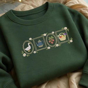 Puede incluir: Sudadera verde oscuro con diseños bordados. Los diseños incluyen un ganso, una tetera azul, fresas y una cesta de frutas, cada uno enmarcado con un borde floral. La sudadera está hecha de un material suave.