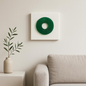 Op de afbeelding: Een modern wandkunstwerk met een levendig groen, cirkelvormig, geplooid ontwerp, gemonteerd op een vierkant wit canvas. Het kunstwerk is een opvallend accentstuk en voegt kleur en textuur toe aan elke interieurruimte. Een uniek woondecoratie-item.