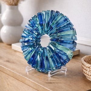 May include: A decorative glass sculpture in a ring shape, featuring shades of blue and teal. The art piece is displayed on a clear stand, showcasing its intricate design. The sculpture is placed on a wooden surface, adding a touch of elegance to any decor.