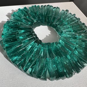 Op de afbeelding: Een cirkelvormige, teal-gekleurde glazen sculptuur met een radiaal ontwerp. De sculptuur bestaat uit talrijke rechthoekige glazen stukken, gerangschikt rond een centrale opening. Het glas heeft een getextureerd uiterlijk en staat op een witte ondergrond.