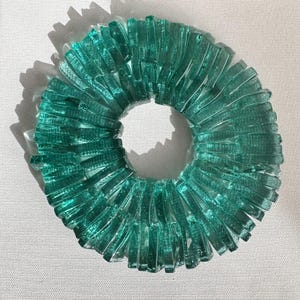 Op de afbeelding: Een cirkelvormig kunstwerk van turquoise glas. Het kunstwerk bestaat uit talrijke rechthoekige glazen stukken die in een cirkelvorm zijn gerangschikt en een ringvorm cre&euml;ren. Het glas heeft een getextureerd uiterlijk en het werk werpt een schaduw.
