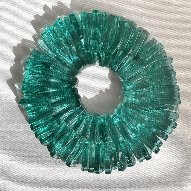Op de afbeelding: Een cirkelvormig kunstwerk van turquoise glas. Het kunstwerk bestaat uit talrijke rechthoekige glazen stukken die in een cirkelvorm zijn gerangschikt en een ringvorm cre&euml;ren. Het glas heeft een getextureerd uiterlijk en het werk werpt een schaduw.