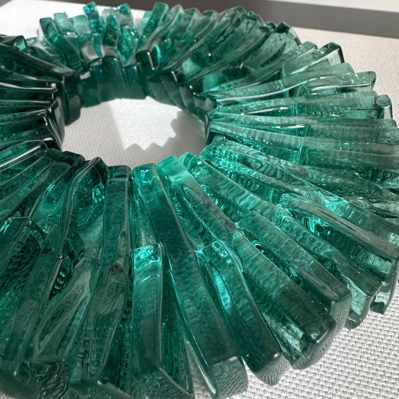 Op de afbeelding: Een cirkelvormig decoratief object gemaakt van teal-kleurige glazen stukken. De glazen stukken zijn rechthoekig en gerangschikt in een radiaal patroon, waardoor een textuurontwerp met een open centrum ontstaat. Het object is waarschijnlijk een decoratieve krans of middelpunt.