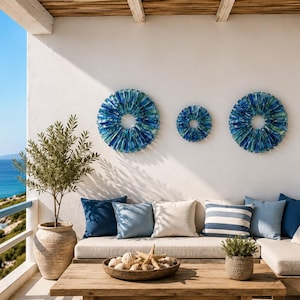 May include: Three circular wall art pieces featuring a radial pattern of blue and teal glass shards. The two larger pieces flank a smaller central piece, all mounted on a white wall above a seating area with blue and white cushions.
