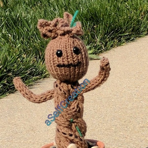 Könnte beinhalten: Eine gestrickte braune Groot-Figur aus der Guardians of the Galaxy-Filmreihe, die in einem Terrakotta-Topf mit kleinen Steinen steht. Die Figur hat ein grünes Blatt auf dem Kopf und lächelt.
