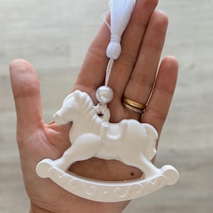 Puede incluir: Adorno blanco de caballo balancín con borla y perla. El adorno se sostiene en una mano, mostrando el diseño del caballo y la borla decorativa. Un recuerdo encantador, ideal para un regalo.