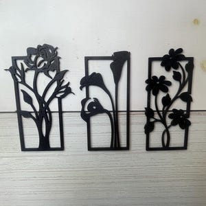 Op de afbeelding: Drie zwarte metalen bloemenmuurkunstwerken. Elk stuk heeft een ander bloemontwerp in een rechthoekig frame. De bloemen zijn onder meer een roos, calla lelies en madeliefjes. De kunst wordt tentoongesteld tegen een witte en houten achtergrond.