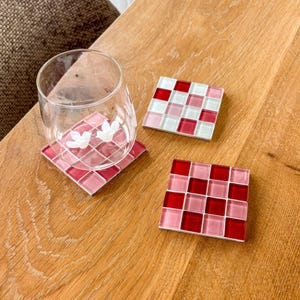 Può includere: Tre sottobicchieri quadrati con un motivo a mosaico nei toni del rosso, rosa e bianco. Un sottobicchiere ha un bicchiere trasparente con un disegno floreale. I sottobicchieri sono su una superficie di legno.