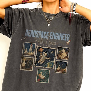 以下が含まれることがあります： 「AEROSPACE ENGINEER Silly Goose」の文字が入った濃いグレーのTシャツ。宇宙にいるガチョウ、ロケット、宇宙飛行士の切手のデザインが特徴です。柔らかい素材で作られたシャツです。