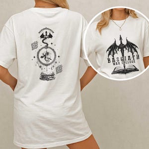 K&ouml;nnte beinhalten: Wei&szlig;es T-Shirt mit einem schwarzen Drachendesign und dem Text "BASGIATH WAR COLLEGE" &uuml;ber einem offenen Buch. Die R&uuml;ckseite des Shirts zeigt ein Drachen-, Mond- und Buchdesign. Das T-Shirt hat eine lockere, &uuml;bergro&szlig;e Passform.