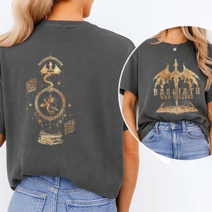 K&ouml;nnte beinhalten: Dunkelgraues T-Shirt mit goldenem Drachendesign. Auf der R&uuml;ckseite befindet sich ein Drache in einem kreisf&ouml;rmigen Rahmen, umgeben von Sternen und B&uuml;chern. Auf der Vorderseite steht "BASGIATH WAR COLLEGE" &uuml;ber einem offenen Buch und einem Drachen.