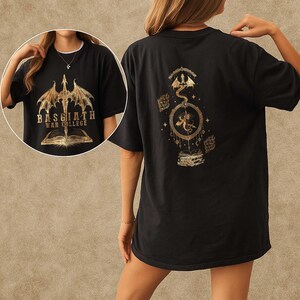 K&ouml;nnte beinhalten: Schwarzes T-Shirt mit goldenen Drachenmotiven auf Vorder- und R&uuml;ckseite. Auf der Vorderseite ist ein Drache und der Text "BASGIATH WAR COLLEGE" &uuml;ber einem offenen Buch abgebildet. Die R&uuml;ckseite zeigt einen Drachen in einem kreisf&ouml;rmigen Rahmen.
