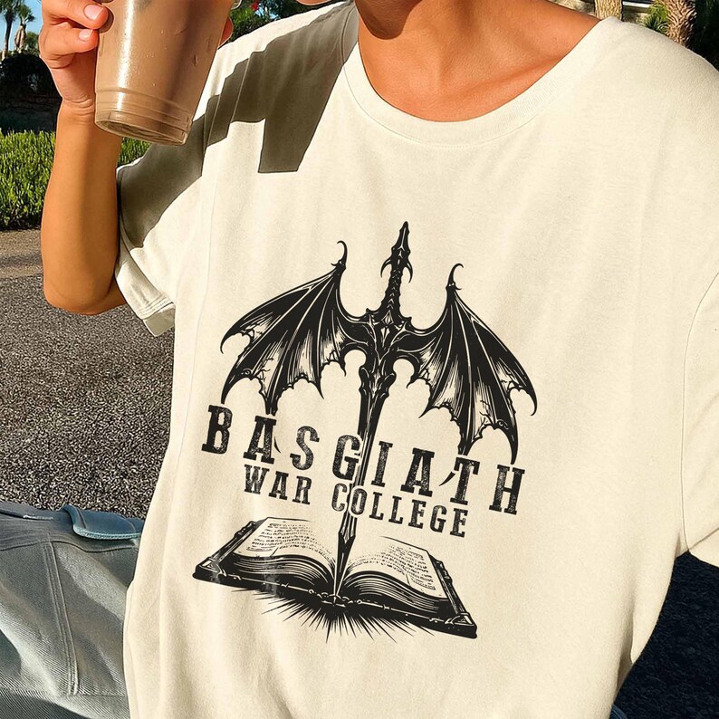 K&ouml;nnte beinhalten: Cremefarbenes T-Shirt mit einer schwarzen Grafik eines Schwertes mit Drachenfl&uuml;geln, das ein offenes Buch durchbohrt. Der Text "BAS GIATH WAR COLLEGE" ist unter dem Schwert- und Buchdesign aufgedruckt.
