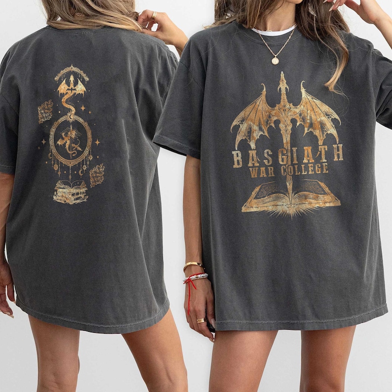 K&ouml;nnte beinhalten: Dunkelgraue T-Shirts mit goldfarbenen Drachen- und Buchgrafiken. Ein Shirt zeigt ein Drachendesign mit dem Text "BASGIATH WAR COLLEGE". Das andere Shirt hat ein Drachen- und Buchdesign.