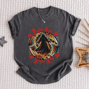Puede incluir: Camiseta gris oscuro con un dise&ntilde;o de una figura encapuchada con espadas, sobre un dise&ntilde;o circular colorido con detalles rojos. El gr&aacute;fico tiene un aspecto vintage y desgastado. Se ve un collar plateado.