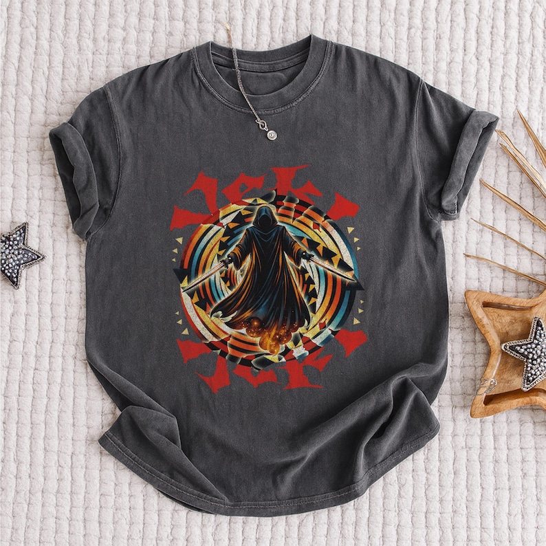 Puede incluir: Camiseta gris oscuro con un dise&ntilde;o de una figura encapuchada con espadas, sobre un dise&ntilde;o circular colorido con detalles rojos. El gr&aacute;fico tiene un aspecto vintage y desgastado. Se ve un collar plateado.