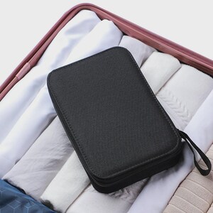 Peut inclure: Un organisateur de voyage rectangulaire noir avec une fermeture &eacute;clair et une petite poign&eacute;e en boucle. L'organisateur est plac&eacute; dans une valise remplie de v&ecirc;tements blancs et gris clair pli&eacute;s.
