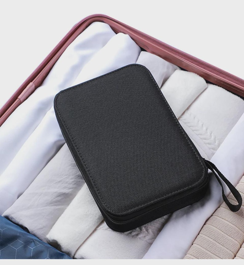 Peut inclure: Un organisateur de voyage rectangulaire noir avec une fermeture &eacute;clair et une petite poign&eacute;e en boucle. L'organisateur est plac&eacute; dans une valise remplie de v&ecirc;tements blancs et gris clair pli&eacute;s.