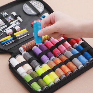 Peut inclure: Une trousse de couture noire zipp&eacute;e contenant des bobines de fil de diff&eacute;rentes couleurs, des aiguilles, des ciseaux et d'autres outils. Une main tient une bobine de fil bleu. La trousse contient du fil rose, violet, orange et vert.