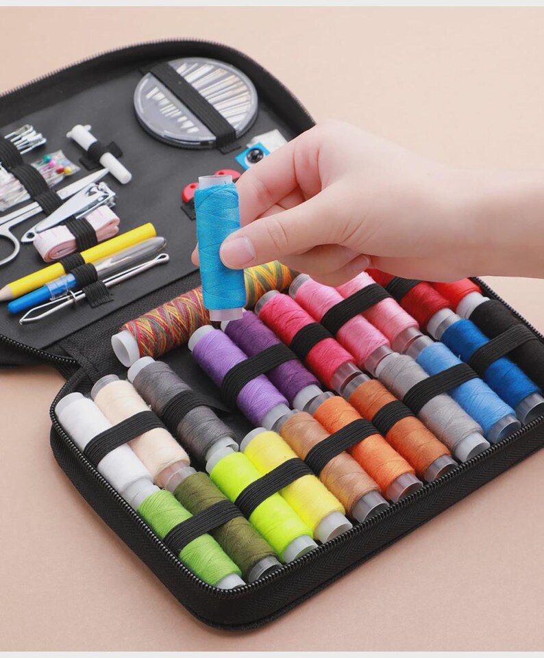 Peut inclure: Une trousse de couture noire zipp&eacute;e contenant des bobines de fil de diff&eacute;rentes couleurs, des aiguilles, des ciseaux et d'autres outils. Une main tient une bobine de fil bleu. La trousse contient du fil rose, violet, orange et vert.
