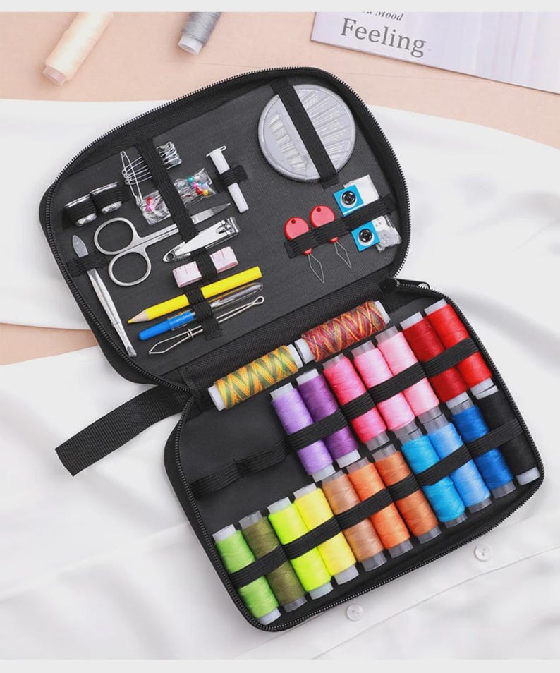 Peut inclure: Trousse de couture noire ouverte, r&eacute;v&eacute;lant divers outils de couture et des bobines de fil color&eacute;es. Le kit comprend des ciseaux, des aiguilles, du fil et d'autres fournitures essentielles pour les projets de couture. Le mot "Feeling" est visible en arri&egrave;re-plan.