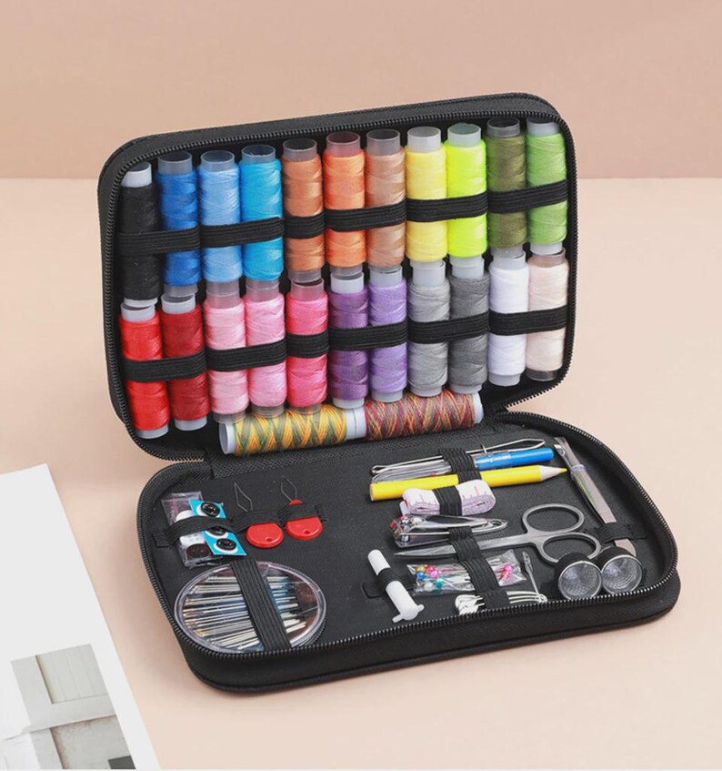 Peut inclure: Trousse de couture noire ouverte, contenant une vari&eacute;t&eacute; de bobines de fil color&eacute;es, des aiguilles, des ciseaux et d'autres outils de couture. Le kit comprend des bobines de fil rouge, rose, bleu, jaune et vert. Le kit est ouvert, r&eacute;v&eacute;lant les fournitures de couture.