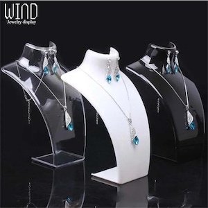 May include: Three jewelry display busts in clear, white, and black. Each bust holds a silver necklace and matching earrings with blue gemstone pendants. The clear bust has a transparent base, while the white and black busts have solid bases. The logo "WIND jewelry display" is in the top left corner.