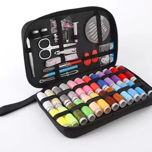 Peut inclure: Trousse de couture noire zipp&eacute;e, ouverte pour r&eacute;v&eacute;ler divers outils de couture et une s&eacute;lection de bobines de fil color&eacute;es. Comprend des ciseaux, des aiguilles, des d&eacute;s &agrave; coudre et un m&egrave;tre ruban.