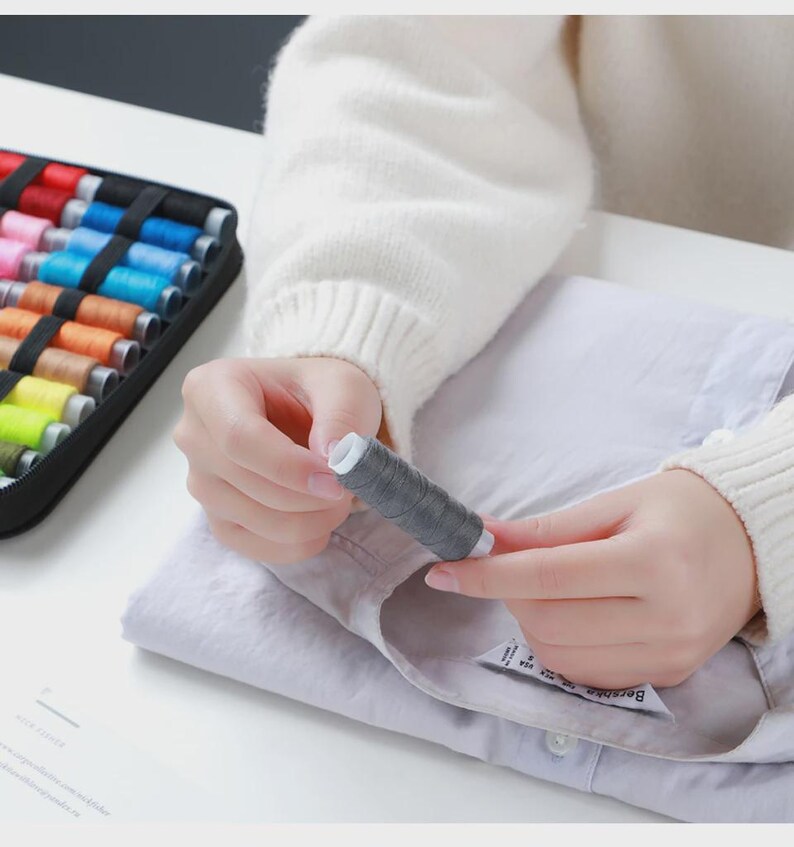 Peut inclure: Une personne tient une bobine de fil gris, avec un c&oelig;ur blanc, au-dessus d'une chemise violet clair. Un &eacute;tui noir rempli de bobines de fil color&eacute;es est visible en arri&egrave;re-plan, sugg&eacute;rant un th&egrave;me de couture ou d'artisanat.