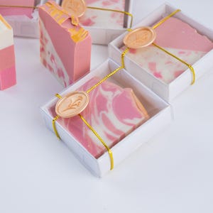 May include: Several bars of handmade soap in white boxes, tied with gold ribbon. The soaps are pink and white, with some having a marbled effect and others a layered design. Each box is sealed with a gold wax stamp.