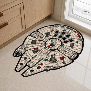 Puede incluir: Una alfombra beige y negra con la forma de la nave espacial Halcón Milenario. La alfombra presenta ilustraciones detalladas en tonos grises, rojos y azules. La alfombra está colocada sobre un suelo de baldosas claras cerca de un mueble de madera y una ventana.