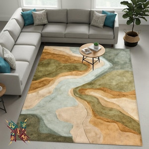 May include: A large area rug with an abstract landscape design. The rug features a river-like pattern in shades of blue and beige, surrounded by brown and green terrain. The rug is placed in a living room with a gray sectional sofa and a small round coffee table.