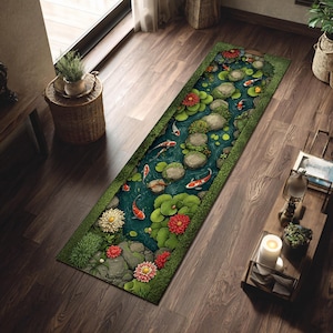 Op de afbeelding: Een rechthoekig tapijt met een vijverscène met koi-vissen, waterlelies en bloemen. Het tapijt heeft een donkerblauw midden met een groene rand en de koi-vissen zijn oranje en wit. De bloemen zijn rood en wit.