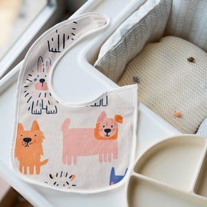 K&ouml;nnte beinhalten: Cremefarbenes Baby-L&auml;tzchen mit bunten Cartoon-Hunde-Illustrationen. Das L&auml;tzchen liegt auf einem wei&szlig;en Hochstuhl-Tablett, neben einem geteilten Teller und einem kleinen Kissen. Das L&auml;tzchen zeigt orange, rosa und blaue Hunde.