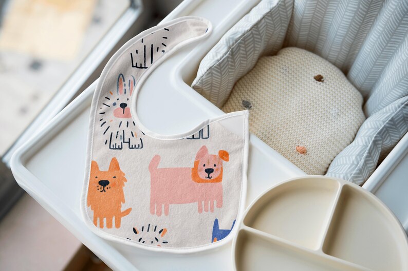 K&ouml;nnte beinhalten: Cremefarbenes Baby-L&auml;tzchen mit bunten Cartoon-Hunde-Illustrationen. Das L&auml;tzchen liegt auf einem wei&szlig;en Hochstuhl-Tablett, neben einem geteilten Teller und einem kleinen Kissen. Das L&auml;tzchen zeigt orange, rosa und blaue Hunde.