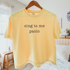 May include: A pale yellow short-sleeve t-shirt featuring the black text "sing to me paolo" printed in a simple, sans-serif font across the center of the chest. The garment has a crew neckline and is displayed on a wooden hanger.