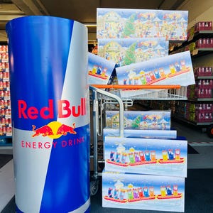 Könnte beinhalten: Ein großer Red Bull Energy-Drink-Display mit blauem und weißem Design, mit dem Red Bull-Logo und den Worten "ENERGY DRINK". Mehrere Adventskalender mit Weihnachtsmotiven sind auf einem Einkaufswagen gestapelt.