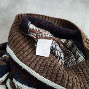 May include: Close-up of a brown knitted jumper with a ribbed collar and patterned details. The jumper features a mix of brown, navy, and white geometric designs. A white tag with text is visible.