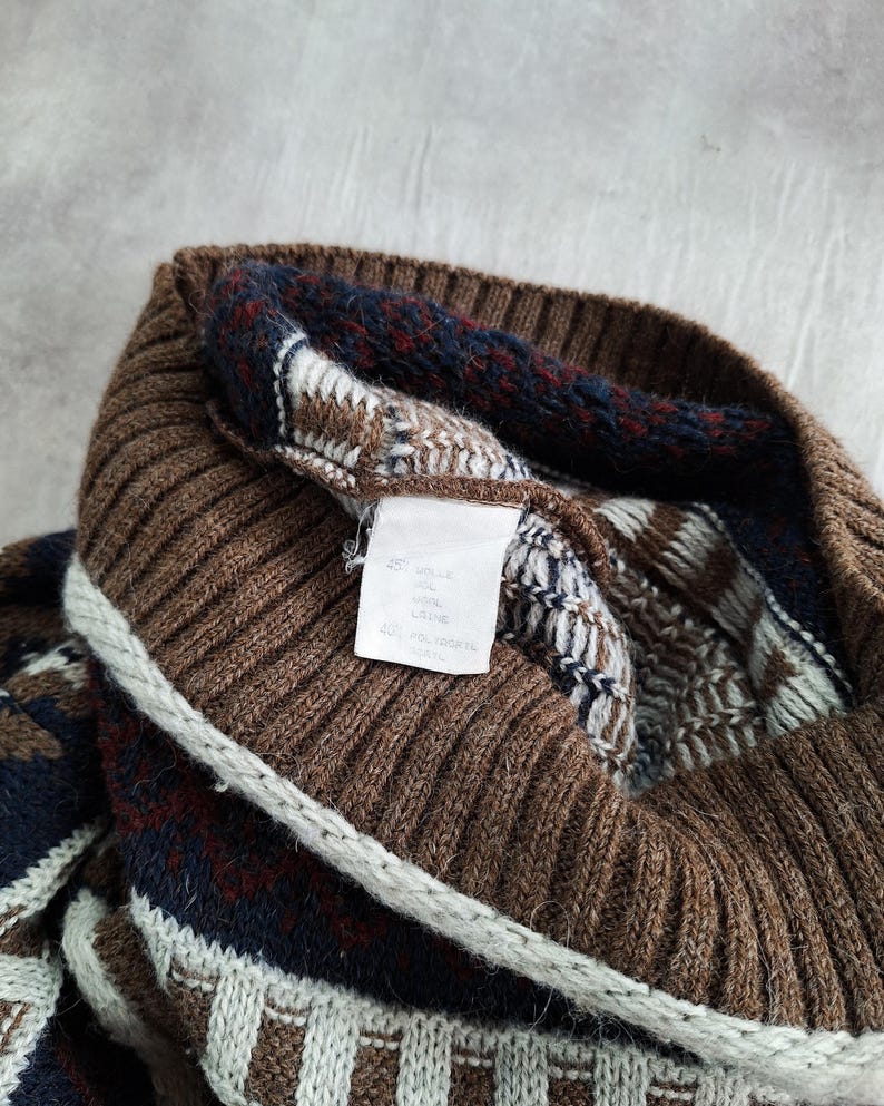 May include: Close-up of a brown knitted jumper with a ribbed collar and patterned details. The jumper features a mix of brown, navy, and white geometric designs. A white tag with text is visible.