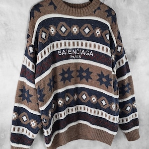 May include: A brown, navy, and white patterned sweater with the text "BALENCIAGA PARIS" embroidered across the chest. The sweater features a crew neck and long sleeves, with a geometric and star design.
