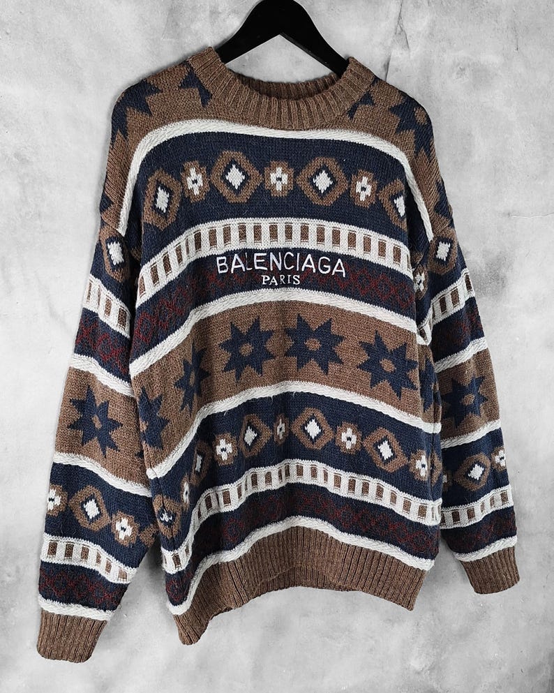 May include: A brown, navy, and white patterned sweater with the text "BALENCIAGA PARIS" embroidered across the chest. The sweater features a crew neck and long sleeves, with a geometric and star design.