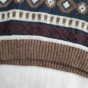 May include: Close-up of a knitted sweater with a geometric pattern. The design features diamonds and floral shapes in brown, white, and navy blue. The bottom portion is a ribbed brown knit.