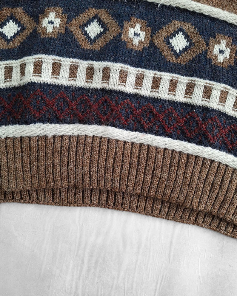 May include: Close-up of a knitted sweater with a geometric pattern. The design features diamonds and floral shapes in brown, white, and navy blue. The bottom portion is a ribbed brown knit.