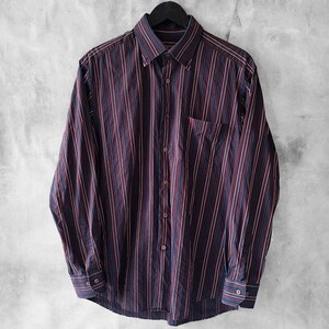 Puede incluir: Camisa de manga larga a rayas verticales en burdeos, azul marino y blanco. La camisa tiene un bolsillo en el pecho y está colgada de una percha negra sobre un fondo gris.