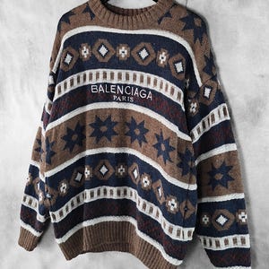 May include: A brown, navy blue, and white patterned sweater with the text "BALENCIAGA PARIS" across the chest. The sweater features a crew neck and long sleeves. The design includes geometric shapes and star patterns.