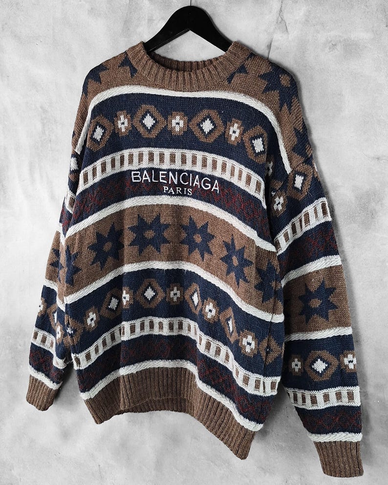 May include: A brown, navy blue, and white patterned sweater with the text "BALENCIAGA PARIS" across the chest. The sweater features a crew neck and long sleeves. The design includes geometric shapes and star patterns.