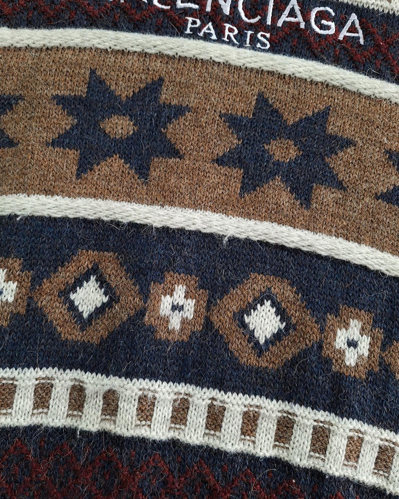May include: Close-up of a knitted sweater with a repeating pattern. The design features brown, navy blue, and white geometric shapes, including stars and diamonds. The text "BALENCIAGA PARIS" is visible at the top.