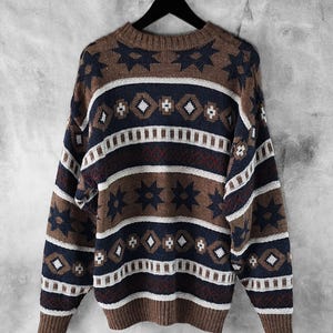 May include: A patterned jumper in brown, navy, and white, hanging on a black hanger. The design features geometric shapes, including stars and diamonds. The colour scheme creates a vintage aesthetic. The jumper is made of a knitted material.