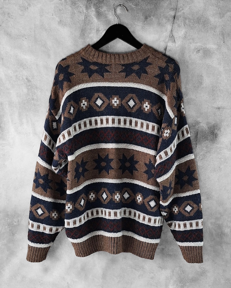 May include: A patterned jumper in brown, navy, and white, hanging on a black hanger. The design features geometric shapes, including stars and diamonds. The colour scheme creates a vintage aesthetic. The jumper is made of a knitted material.