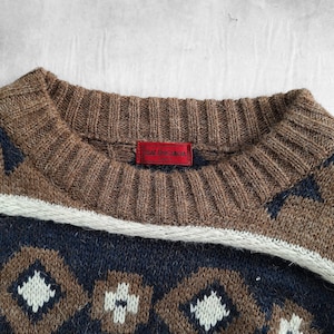 May include: Close-up of a brown, navy, and white patterned sweater. The sweater has a ribbed brown collar and a red tag with the brand name "BALENCIAGA". The design includes geometric shapes and a white stripe.
